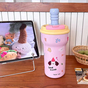 Pink children's cup with cartoon designs on a wooden table next to a tablet displaying cartoon characters.