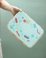Colorful geometric patterned lunch box held by a hand against a green background