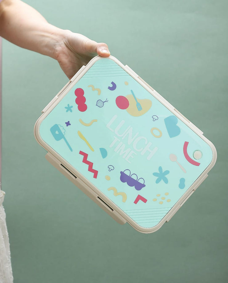 Colorful geometric patterned lunch box held by a hand against a green background
