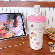White cup with pink lid featuring cartoon designs on a wooden surface next to a tablet displaying plush toys.